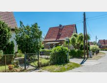 Einfamilienhaus mit rotem Satteldach in einer ländlichen Wohngegend, umgeben von einem Garten mit Bäumen und Sträuchern, sowie einem Maschendrahtzaun entlang der Straße. Straßenschilder mit den Bezeichnungen „Heineallee“ und „Lönsweg“ sind an einer Ecke gut sichtbar, im Hintergrund weitere Häuser.