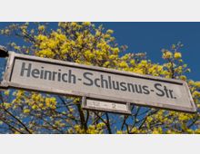 Straßenschild mit der Aufschrift „Heinrich-Schlusnus-Str.“, im Hintergrund grüne Baumkronen und blauer Himmel.