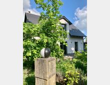 Modernes Einfamilienhaus mit weißer Fassade und Schrägdach aus dunklen Ziegeln, umgeben von grüner Bepflanzung. Im Vordergrund steht eine Steinstele mit einer schwarz-weißen Kugel darauf.