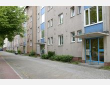 Wohnblock in einer städtischen Straße mit vierstöckigen Gebäuden aus Putzfassaden in Beige- und Blautönen. Vor den Gebäuden befinden sich gepflasterte Gehwege, niedrige Büsche und ein Baum; einige Haustüren haben Vordächer aus Glas.