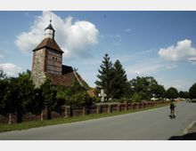 Historische Dorfkirche mit Fachwerk- und Backsteinelementen, umgeben von Bäumen und einer niedrigen Backsteinmauer, mit einem Fahrradfahrer auf einer angrenzenden Straße unter blauem Himmel.