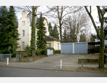 Gelbes Wohngebäude im Jugendstil mit geschwungenem Dachgiebel, umgeben von Bäumen und Sträuchern, rechts davon eine Reihe von blauen Garagentoren; davor gepflasterte Einfahrt und Bürgersteig.