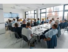 Ein moderner Seminarraum mit großen Fenstern, durch die die Oberbaumbrücke und ein Teil der Berliner Skyline sichtbar sind. Eine Gruppe von Personen sitzt an langen Tischen und hört einem Vortragenden zu, der vor zwei großen Bildschirmen steht.