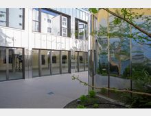 Moderner Innenhof mit spiegelnder Glasfassade, großen bodentiefen Fenstern und begrünten Elementen. Der Boden besteht aus glatter, gepflasterter Fläche, während eine Pflanze im Vordergrund zu sehen ist.