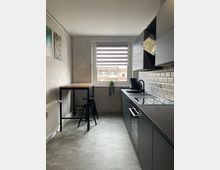 Moderne, schmale Küche mit grauen Schränken und schwarzer Arbeitsfläche, einem kleinen Esstisch mit zwei schwarzen Hockern und einer Fensterfront mit gestreifter Jalousie; die Wände teilweise in Betonoptik, eine weiße Fliesenrückwand und helle Bodenfliesen.