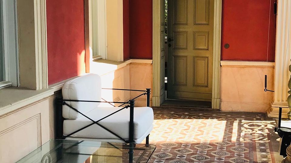 Überdachter Korridor mit roten Wänden und hellen Stuckrahmen, einem alten Holzportal am Ende und einem dekorativen Fliesenboden mit geometrischem Muster. Entlang der Fensterfront stehen weiß gepolsterte Sitzmöbel und ein gläserner Couchtisch mit Metallgestell.