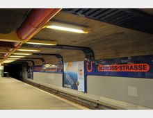 U-Bahnhof mit dem Namen „Schloßstraße“, erkennbar durch blaue Wände mit orangener Beschriftung und U-Bahn-Logos. Die Decke besteht aus Beton, und eine beleuchtete Plattform erstreckt sich entlang der Gleise.