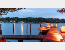 Eine Abendaufnahme eines Sees mit einem beleuchteten hölzernen Steg, an dem ein kleines Motorboot angedockt ist. Im Hintergrund sind eine Baumreihe und ein Hafen mit mehreren Segelbooten sowie beleuchtete Gebäude zu sehen.