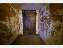 Ein schmaler Kellerraum mit abblätternder Wandfarbe führt zu einer schweren Metalltür, auf der Aufschriften wie „Männer Toiletten Frauen“ und „Bei Umluftbetrieb öffnen“ zu erkennen sind. Der Boden ist mit grauen Fliesen bedeckt, und die Atmosphäre wirkt spartanisch und funktional.