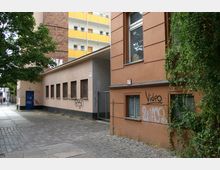 Eingangsbereich eines Gebäudekomplexes in einer urbanen Umgebung, mit einer beigen Fassade, einer blauen Tür und mehreren vergitterten Fenstern, die mit Graffiti versehen sind. Im Vordergrund sind gepflasterte Gehwege und ein kleiner Baum zu sehen, während im Hintergrund ein mehrstöckiges Wohnhaus mit gelben Balkonen erkennbar ist.