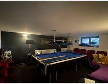 Ein gemütlich eingerichteter Billardraum mit einem Billardtisch in der Mitte, blauem Tuch und Hängelampen mit blauen Schirmen darüber. Im Hintergrund befinden sich rote Kinosessel, ein Sofa mit goldfarbenen Kissen, eine Bar-Ecke sowie ein kleines Fenster mit Blick nach draußen.