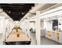 Ein moderner, offener Arbeitsbereich mit langen Holztischen, schwarzen Stühlen und weißen Säulen. Im Hintergrund befinden sich Wanddekorationen, während rechts eine kleine Kaffeebar mit Regalen und einer Theke sichtbar ist.