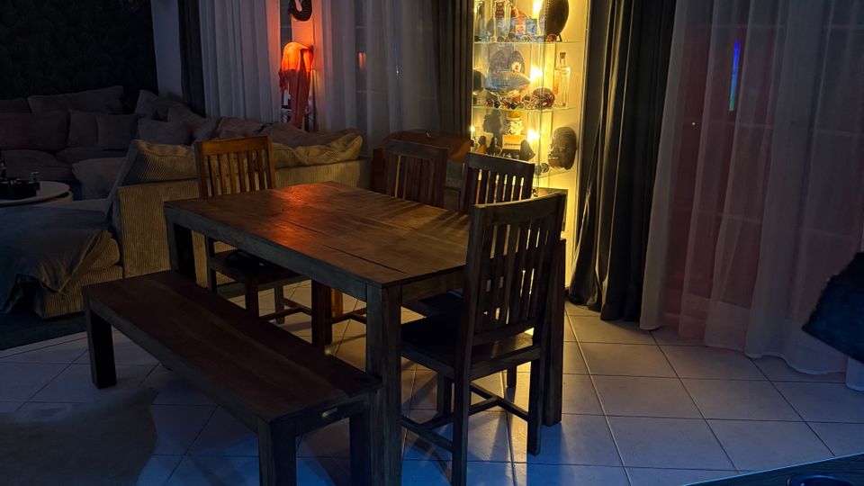 Wohn- und Essbereich mit Holz-Esstisch, Stühlen und Sitzbank auf hellem Fliesenboden, daneben ein graues Sofa und ein beleuchteter Glasvitrinenschrank. Große bodentiefe Fenster mit weißen Gardinen und dunklen Vorhängen säumen die Wand; im Vordergrund ist der Rand eines Billardtischs zu sehen.