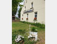 Eine beige Fassade eines ländlichen Gebäudes mit dem Schild „Zum Heiratsmarkt“ über den Fenstern, die mit roten Geranien dekoriert sind. Im Vordergrund befindet sich ein kleiner Garten mit einem weißen, dekorativen Wagenrad und blühenden Blumen.