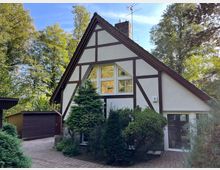 Fachwerkhaus mit weißer Fassade und braunen Holzbalken, steilem Satteldach und einem großen dreigeteilten Fenster im Giebel. Umgeben von Bäumen, Büschen und einem gepflasterten Weg, im Hintergrund eine Garage aus Holz.