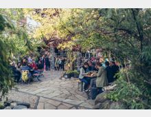 Ein Außenbereich mit vielen Menschen, die an Holztischen auf Steinplatten sitzen und speisen. Umgeben von Bäumen und Pflanzen, während sich im Hintergrund weitere Gruppen unter einer offenen Konstruktion aus Metallträgern versammeln.