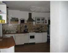 Küche mit weißen Schränken, einer Dunstabzugshaube, und einem Herd. Im Hintergrund befinden sich beige Wandfliesen, ein Spülbecken und Küchenutensilien; der Boden ist aus Holz.