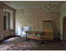 Ein verlassener, heruntergekommener Raum mit beschädigten Wänden und einem Holzfußboden, der sichtbare Abnutzung zeigt. In der Mitte steht eine alte Werkbank, an der Wand lehnen Holztüren und Metallplatten, und von der Decke hängt eine mehrarmige Lampe mit Glasleuchten.