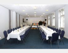 Ein rechteckiger Veranstaltungsraum mit blauem Teppichboden, zwei langen, weiß gedeckten Tischen mit Kerzenständern und blauen Stühlen. Im Hintergrund befinden sich Lautsprecher, ein Tisch mit Dekorationen und ein Klavier.