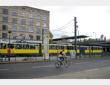Straßenbahn und Fahrradfahrer vor der Haltestelle "Greifswalder Straße" in einer städtischen Umgebung. Im Hintergrund ein mehrstöckiges Gebäude mit zahlreichen Fenstern und ein S-Bahn-Symbol, das auf die Nähe eines S-Bahnhofs hinweist.