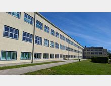 Langes, mehrstöckiges Gebäudetrakt mit heller Putzfassade und Reihen gleichförmiger Fenster, aufgenommen aus schräger Perspektive. Davor verlaufen ein gepflasterter Weg und eine Grünfläche; im Hintergrund ist ein weiterer Gebäudeflügel unter blauem Himmel zu sehen.