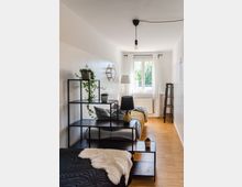 Modern eingerichtetes Schlafzimmer mit Holzboden, einem Fenster mit weißen Vorhängen und Tageslicht. Das Zimmer enthält ein schwarzes Regal mit Pflanzen und Deko-Objekten, zwei Betten mit dekorativen Kissen und Decken, sowie eine Stehlampe und Heizkörper unter dem Fenster.