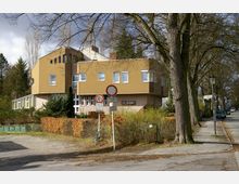 Modernes Gebäude mit gelbbrauner Fassade und großen rechteckigen Fenstern, umgeben von Bäumen, Sträuchern und Hecken. Im Vordergrund eine Straße mit Verkehrsschildern und einem Fußgängerweg sowie eine Straße mit parkenden Autos.