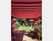 Gartenbereich mit einer rot-weiß gestreiften Markise, zwei roten Liegestühlen auf einer Rasenfläche, einem kleinen Holztisch mit Laterne und roten Blumen in Pflanzgefäßen. Im Hintergrund stehen Bäume, eine Holzlaube und ein Sonnenschirm.