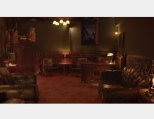 Ein gemütlicher Raum im Vintage-Stil mit dunkelgrünen Wänden und Holzvertäfelung. Der Raum ist mit schwarzen Ledersofas, einem holzernen Schreibtisch, Ledersesseln und einer Hängelampe ausgestattet; im Hintergrund sind ein Fenster, Bücherregale und Trommeln sichtbar.