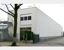 Modernes, einstöckiges Gebäude mit weißer Fassade, großen Fenstern im Erdgeschoss und Metalltreppe an der Seite. Im Vordergrund ein Zaun mit Tor, ein Baum und ein gepflasterter Gehweg.