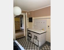 Eine ältere Küche mit gefliesten Wänden in beige und blauen Elementen, einem weißen, gefliesten Holzofen mit schwarzen Metallgriffen und Schubfächern sowie einem Schachbrettmuster-Boden in Grau und Weiß. Eine Tür führt in einen angrenzenden Raum mit gelb gemusterten Wänden und Fliesenboden.