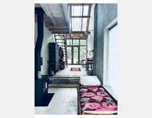 Innenraum eines modernen, loftartigen Wohnhauses mit Betonboden, Holzdeckenbalken und einem großen Oberlicht über dem Durchgang. Im Hintergrund führen eine offene Treppe und hohe Glastüren nach draußen; entlang der Wände stehen ein schwarzer Ofen, ein Spind, ein Arbeitsplatz sowie eine Liege mit gemustertem Teppich neben einem großen Fenster mit Vorhang.