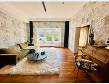 Wohnzimmer mit Holzboden, einer gemusterten Tapete in grauen Tönen und einem großen Fenster mit Blick ins Freie. Der Raum ist mit einem grünen Samtsofa, einem flauschigen weißen Teppich und einem ovalen Couchtisch eingerichtet; im Vordergrund steht ein antiker Holzschreibtisch mit dekorativen Schnitzereien.