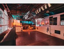 Große, industrielle Halle mit hohen Decken, sichtbaren Stahlträgern und einer Mischung aus roten und blauen Lichtakzenten. An den Wänden befinden sich Fensterfronten und weiße, modulare Strukturen mit einem gemalten Astronautenmotiv, ergänzt durch technisches Equipment und eine auffällige Rohrkonstruktion im Hintergrund.