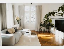 Helles Wohnzimmer mit großen Fenstern und beigen Vorhängen, einem grauen Ecksofa mit Kissen, einem weißen Beistelltisch und mehreren Zimmerpflanzen. Der Raum hat einen Holzboden, einen flachen Fernseher an der Wand und viel Tageslicht.