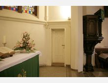 Innenraum einer Kirche mit einem Altar im Vordergrund, der mit einer Bibel, einer Kerze und einem Blumenarrangement dekoriert ist. Im Hintergrund eine helle Tür, verziertes Kirchenfenster und rechts eine dunkle Holzkanzel mit Schnitzereien.