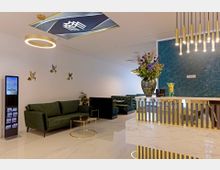 Eleganter Warteraum mit modernen Möbeln, darunter grüne Sofas, ein runder Glastisch mit goldenem Gestell und eine dekorative Empfangstheke mit Marmoroptik. An der Rückwand ist ein grünes Muster mit goldenen Lampen zu sehen, ergänzt durch Blumenarrangements und dekorative Akzente.