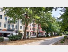 Straßenszene mit Gehweg und Baumreihe im Vordergrund, dahinter mehrstöckige Wohnhäuser mit Balkonen und Erdgeschossgeschäften, darunter eine Pizzeria und ein kleiner Supermarkt; parkende Fahrzeuge und Fußgänger entlang der Straße.