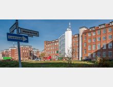 Straßenszene mit roten Backsteingebäuden im Hintergrund, darunter ein modernes Gebäude mit einer großen, weiß verglasten Fassade. Im Vordergrund befinden sich Straßenschilder, darunter eines mit der Aufschrift „Einbahnstraße“, und eine Grünfläche.