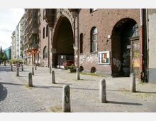 Straßenszene mit historischem Backsteingebäude, das durch bogenförmige Eingänge und Fenster mit weißen Sprossen gekennzeichnet ist; die Fassade ist mit Graffiti versehen. Links stehen moderne Wohnhäuser und Baugerüste, während der gepflasterte Gehweg mit Pollern gesäumt ist.