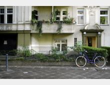 Alt-Text: Wohngebäude mit heller Fassade und dekorativen Stuckelementen. Vor dem Gebäude stehen ein Fahrrad sowie kleinere Pflanzen hinter einem niedrigen Zaun.