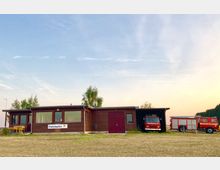 Gebäude einer Feuerwehrstation in Fehrbellin mit Holzfassade, großen Fenstern und einem überdachten Bereich, in dem ein Feuerwehrfahrzeug steht. Im Vordergrund ist ein freies Feld sichtbar, während der Himmel im Hintergrund in Abendfarben leuchtet.