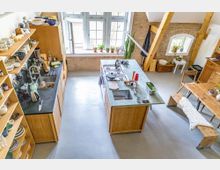 Helle, offene Küche in einem Loft mit Holzbalken, Ziegelwänden und großen Fenstern. Ein zentraler Küchenblock mit Spüle und Herd ist von Holzschränken und Regalen umgeben; im Hintergrund ein Essbereich mit Holztisch und Bank.