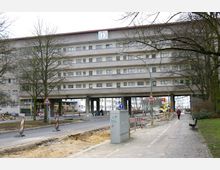 Wohngebäude mit flachem Dach und mehreren horizontalen Fensterreihen, über einer Straße auf Pfeilern gebaut. Im Vordergrund sind Baustellenabsperrungen, ein Gehweg, und Bäume.