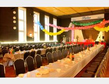 Ein Festsaal mit langen, gedeckten Tischen, an denen schwarze Stühle stehen. Die Decke ist mit bunten Luftballon-Girlanden dekoriert, und im Hintergrund befindet sich eine kleine Bühne mit weiteren Dekorationen.