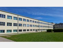 Langer, dreigeschossiger Gebäudetrakt mit heller Fassade und vielen rechteckigen Fenstern in Reihen, davor eine große Rasenfläche und ein Gehweg. Rechts schließen weitere Gebäudeteile sowie geschnittene Hecken an, darüber ein klarer blauer Himmel.
