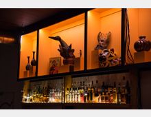 Ein Regal in einer Bar mit beleuchteten Fächern: Oben sind verschiedene dekorative Objekte wie Keramikvasen, Masken, eine Ikone und Figuren ausgestellt. Darunter befindet sich eine beleuchtete Sammlung von Spirituosenflaschen.