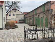 Ein gepflasterter Innenhof mit historischen Backsteingebäuden, die grüne Türen und Fensterläden haben. Links steht ein hell verputztes Wohngebäude, im Hintergrund sind Büsche und ein Baum zu sehen.
