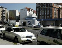 Straßenszene mit mehreren geparkten Taxis im Vordergrund und der U-Bahn-Station „U-Bhf Bayerischer Platz“ im Hintergrund. Umgeben von Wohngebäuden mit Klinker- und hellen Fassaden sowie einem vorbeifahrenden Bus auf der Straße.