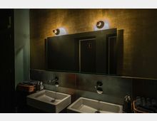 Modern gestaltetes Badezimmer mit zwei weißen Waschbecken und wandmontierten Wasserhähnen. Oberhalb der Waschbecken befindet sich ein großer Spiegel, der von zwei Wandlampen beleuchtet wird, und auf der Ablage stehen Seifenspender neben Körben mit dunklen Handtüchern.